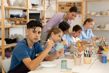 Positive teenage boy painting handmade ceramic cup with brush in pottery workshop