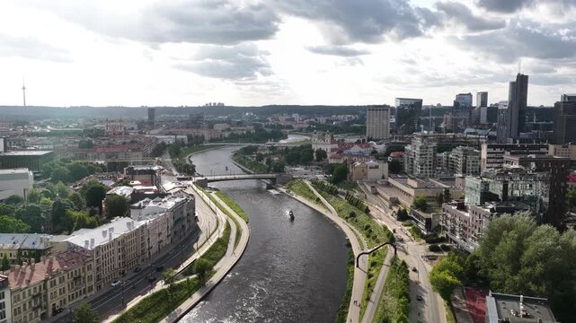 Wide aerial shot of Vilnius, Lithuania, capturing the Neris River, a blend of historic and modern buildings, green spaces, and city landmarks under a cloudy sky.