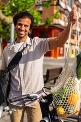 Happy indian businessman holding a reusable shopping bag with groceries hanging from his bicycle...