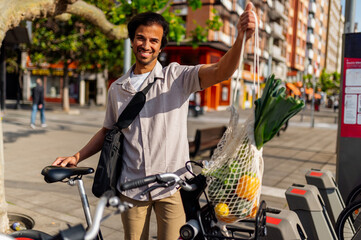 Happy businessman holding reusable shopping bag with groceries while using a bike sharing service...