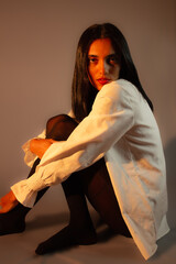 Beautiful woman in white shirt and black tights sitting on the floor under dramatic light. Ideal...