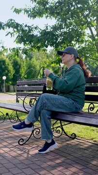 Sporty woman in sunglasses resting on park bench, taking water bottle from backpack and drinking. Healthy lifestyle and outdoor wellness in 4K. vertical video