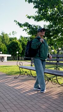 Sporty woman wearing sunglasses and baseball cap walks to park bench, sits down, waves, talks on phone and shows video. Casual urban lifestyle in 4K.