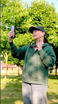 Sporty smiling woman in sunglasses and baseball cap flirting playfully at smartphone camera, enjoying sunny day. 4K lifestyle footage. vertical frame.