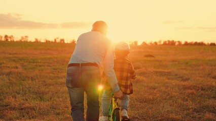 Dad helps his son ride bike. Boy kid, dad play together, sunset. Father teaches child son wearing safety helmet to ride bicycle. Family in park. Child rides bicycle. Child dream learns to ride bicycle