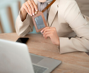 A woman's body wearing a hijab and formal attire holds an empty ID card holder in front of a laptop.