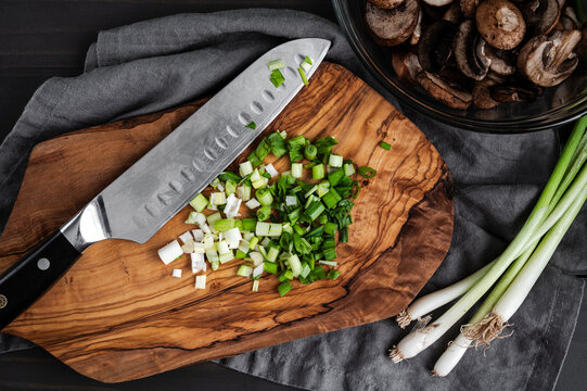 Sliced Scallions on a Wooden Cutting Board: Sliced green onions on a chopping board with a santoku knife and sliced cremini mushrooms on the side