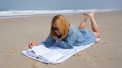A young woman lies on her stomach on a beach towel, reading a book by the Atlantic Ocean. She enjoys a peaceful, sunny day with waves crashing gently in the background