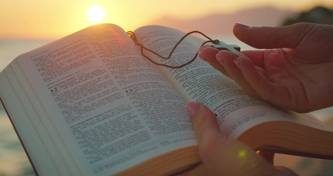 Hands hold open Bible book and wooden cross at beach during glowing sunset