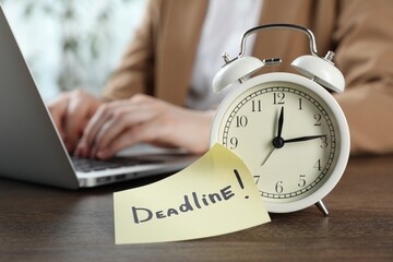 Deadline. Woman with laptop working at wooden table in office, focus on alarm clock and sticky note