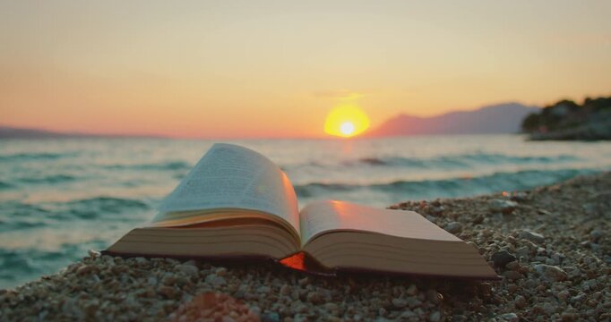Bible pages turning on pebble beach at sea with glowing sunset light