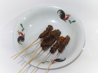Six skewers of grilled meat on a rooster-patterned plate against a clean white background.