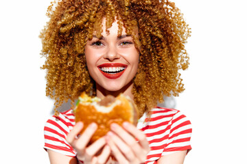 Cheerful young woman with curly hair enjoying a delicious burger, wearing a striped shirt, against...