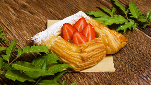 Freshly baked puff pastry filled with sliced strawberries and apricot jam, sprinkled with powdered sugar, lying on a wooden table near some green leaves