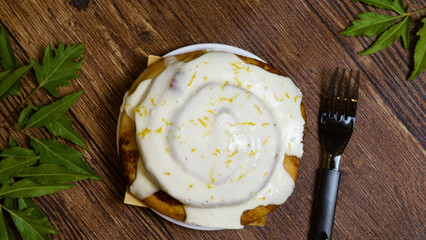 Freshly baked cinnamon roll topped with a generous layer of lemon frosting sits on a wooden table, ready to be enjoyed, garnished with lemon zest and accompanied by a fork