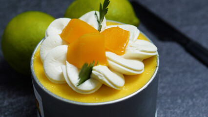 Small mango mousse cake decorated with whipped cream rosettes, mango slices, and fresh herbs, sitting on a dark gray surface with whole limes in the background