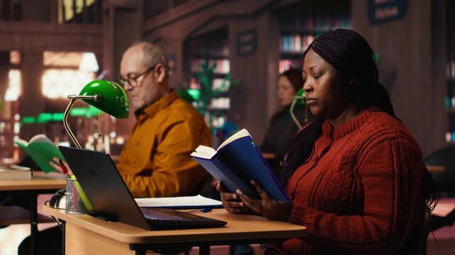 Black female student taking notes surrounded by books and study materials, preparing for a master dissertation and researching academic resources at the campus library. Camera B.