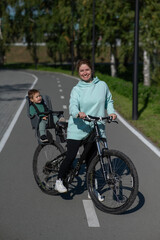 Caucasian woman riding bicycle with toddler son sitting behind her in child seat in park. Vertical...