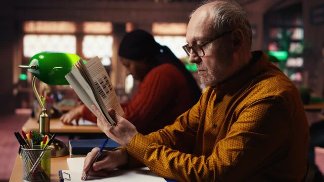 Senior male student reading a book in a study area at a public library, reviewing papers and looking for thesis citations sources. Aged man determined to graduate college. Camera B.