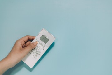 Hand holding a white AC remote, fingers on buttons, against light blue. Ideal for climate control, comfort, technology, and home appliances.