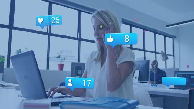 Mature woman typing at office desk and monitoring animated social media icons rising above laptop