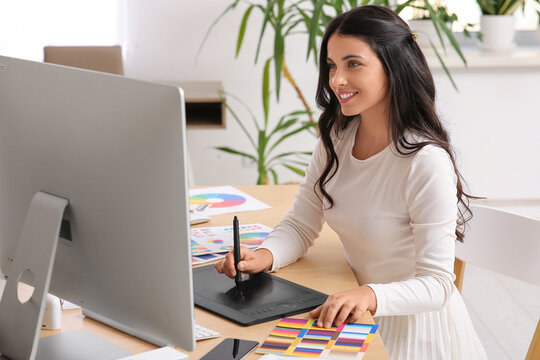 Young female graphic designer working with tablet, color palettes and computer at table in office - Powered by Adobe