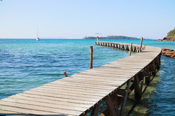 Curved wooden pier extending into turquoise waters with a distant yacht and island backdrop in Klong Mad fisherman village, Koh Kood, Thailand.