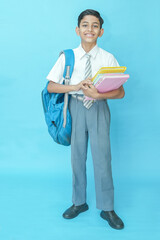 Happy Indian student schoolboy wearing uniform holding books and bag standing isolated over blue...