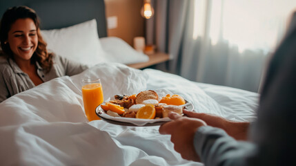 A man is holding breakfast in bed for his wife, who smiles and looks at him with joy as she sits up from the bed.