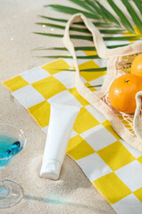 Flat lay tropical summer setup with tropical leaves, cosmetics tube, oranges, and cocktail on checkered towel. Soft sand and bright sun create an ideal scene for design or beauty product mockup.