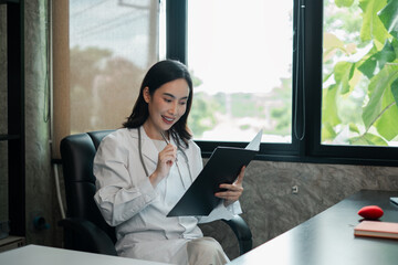 A professional woman in a white coat reviews documents in a modern office with large windows and...