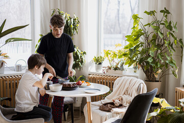 Two brothers having breakfast in sunny room with houseplants, cake, fruit, and cozy cat