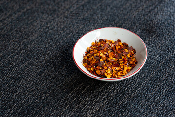 Red chili flakes, red pepper flakes, in a white porcelain sauce cup, placed on grey fabric napkin. In cozy home environment