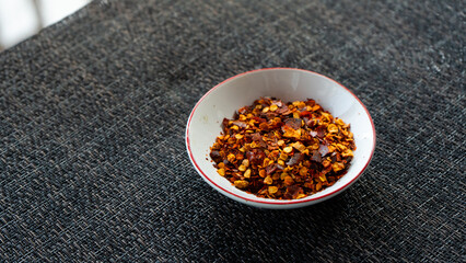 Red chili flakes, red pepper flakes, in a white porcelain sauce cup, placed on grey fabric napkin. In cozy home environment