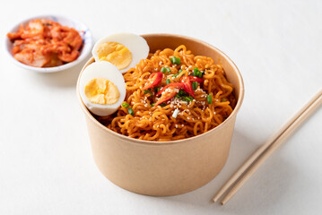 Spicy Korean dried instant noodles with boiled egg, sliced scallions, sesame seed and fresh chili in paper bowl on white background