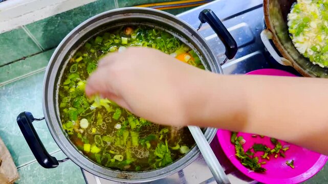 Traditional Indonesian beef soup cooked in a pot on a gas stove. A woman hand adds seasonings such as sliced scallions and fried onions. Top view.
