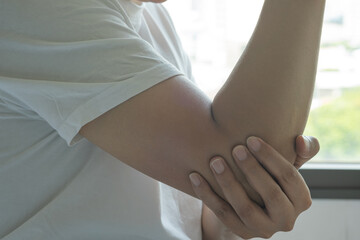 An Asian man grasping a swollen elbow indoors near a window, suggesting soft tissue injury or...