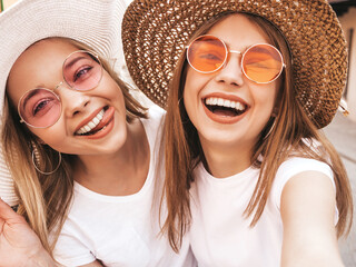 Two young smiling hipster blond women in summer white t-shirt. Female taking selfie self portrait...