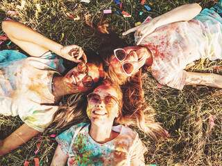 Three happy beautiful female making party at Holi colors festival in summer time.Young  smiling women friends having fun after music event at sunset.Models lying on the grass in sunglasses