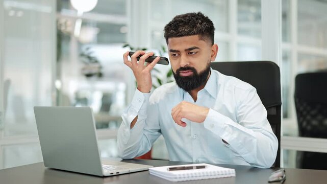 Indian professional businessman, dressed in a light blue shirt, intently listens to a voice message on his smartphone at a desk in a modern office, showing a focused expression.