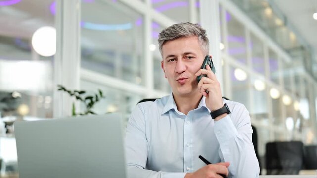 Confident businessman in a light blue shirt happily talks on a mobile phone, working on a laptop in a bright modern office, demonstrating effective communication and concentration.