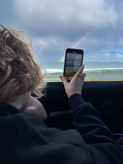 Teenage boy taking photo of rainbow over ocean on phone from car