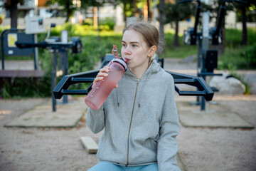 Young woman drinking water after workout at outdoor gym in summer park