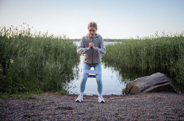 woman doing resistance band squat exercise near lake in summer