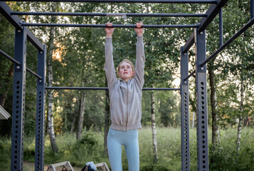 Young woman hanging on pull-up bar at outdoor gym in park
