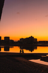 Two people silhouetted at sunset on coastal path near city skyline