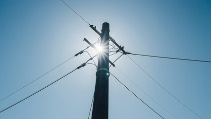 Fototapeta premium Sun shines brightly behind a tall utility pole carrying power lines against a clear blue sky.