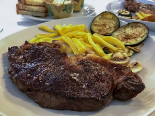 portion of entrecote with french fries and grilled vegetables, on a white plate served at the table in a restaurant