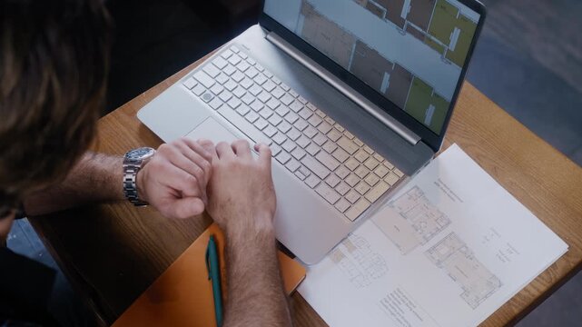 A stylish man analyzes architectural plans on a laptop while making handwritten notes in a notebook at a cafe table.