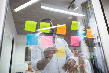 Smiling manager sticking colorful adhesive notes on a glass wall, sharing ideas during a...
